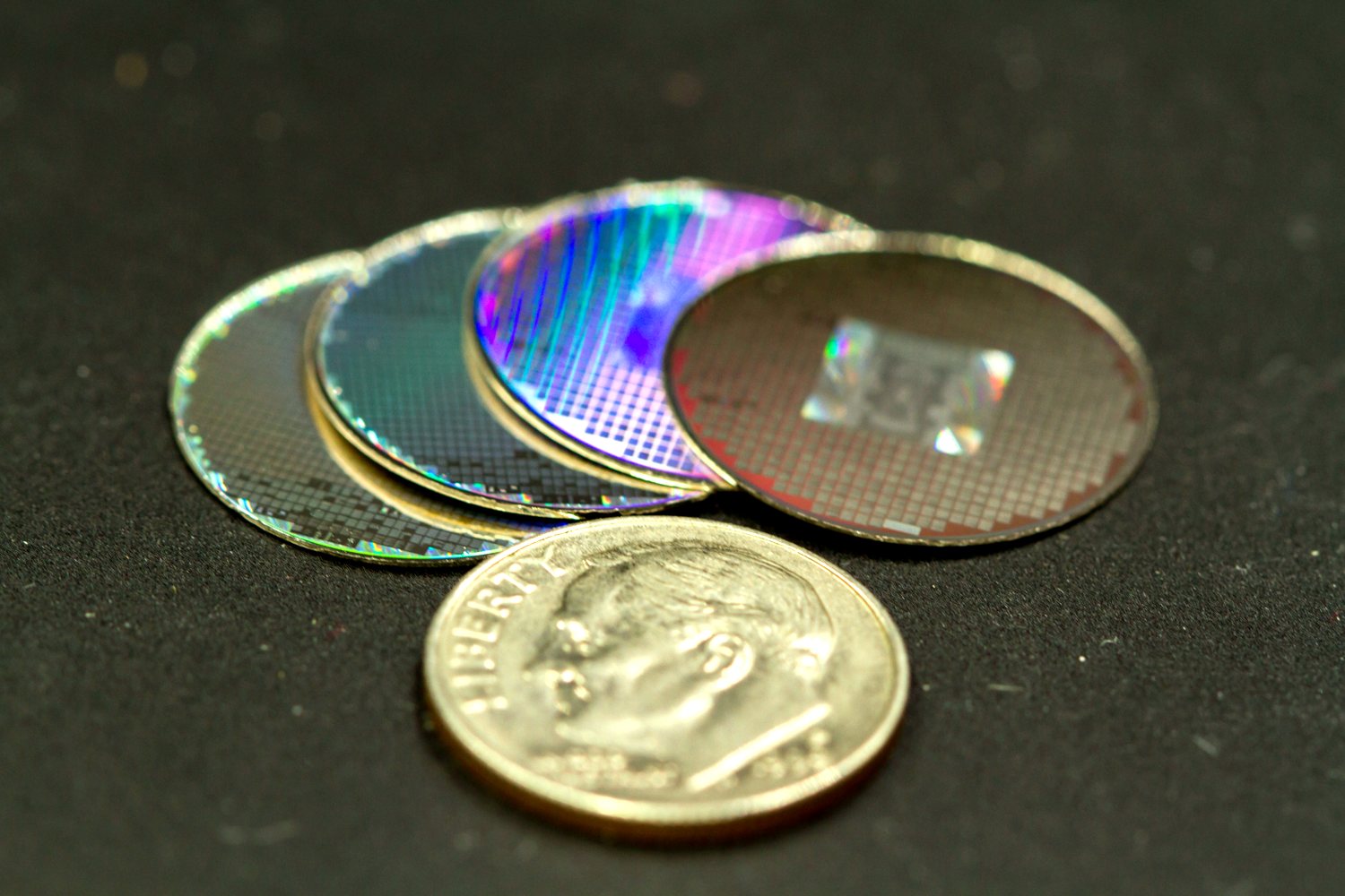 Four NanoFiche nickel discs fanned out, their engraved micro-grids catching the light in iridescent color, with a United States dime resting in front of them for scale.
